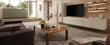 Wohnzimmer mit weißer Wohnwand, großem Fernseher, Holzcouchtisch und grauem Teppich vor hoher Fensterfront.